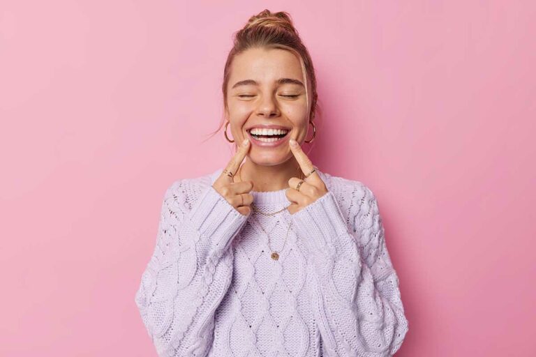 young-glad-woman-points-toothy-smile-shows-perfect-white-teeth-being-good-mood-keeps-eyes-closed-dressed-casual-knitted-jumper-isolated-pink-background-positive-emotions-concept